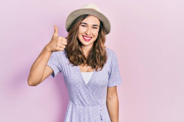Young hispanic girl wearing summer hat smiling happy and positive, thumb up doing excellent and approval sign 