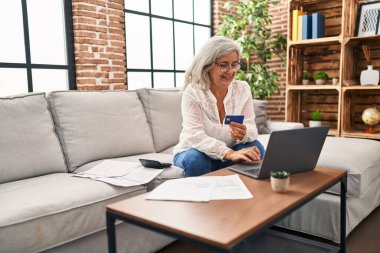 Evde dizüstü bilgisayar ve kredi kartı kullanan orta yaşlı kadın.