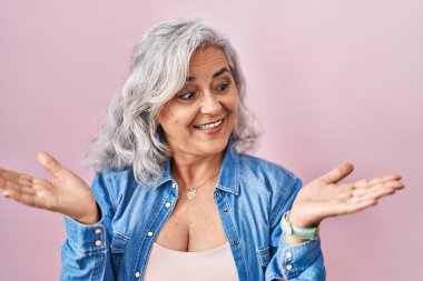 Pembe arka planda duran gri saçlı orta yaşlı kadın avuçlarını açıp gülümsüyor, sunuyor ve reklam karşılaştırması yapıyor ve dengeyi sağlıyor. 