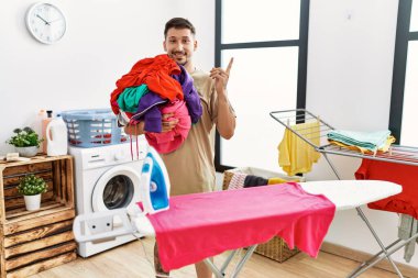 Young handsome man holding laundry ready to iron smiling happy pointing with hand and finger to the side 