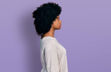 Afro-Amerikalı genç bir kadın, günlük giysiler içinde rahat bir görünüm takınıyor, doğal bir yüz ifadesi ve kendine güvenen bir gülümsemeyle poz veriyor.. 