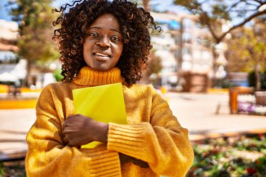 Afro-Amerikalı kadın gülümsüyor. Parkta kitap tutuyor.