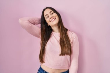 Genç esmer kadın pembe arka planda duruyor gülümsüyor saçlarına dokunuyor, el hareketi yapıyor, çekici ve şık duruyor. 