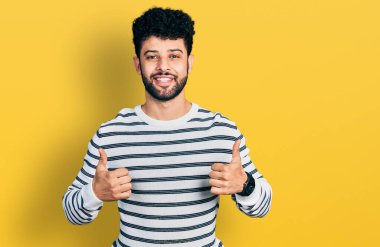 Sakallı, sıradan çizgili süveter giyen, el hareketleriyle olumlu jest yapan, gülümseyen ve mutlu genç bir Arap. Neşeli ifade ve kazanan jest. 