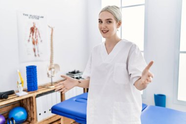 Ağrı giderme kliniğinde çalışan beyaz bir kadın. Kollarını açıp gülümsüyor. Dostça karşılama, olumlu ve kendinden emin selamlar. 