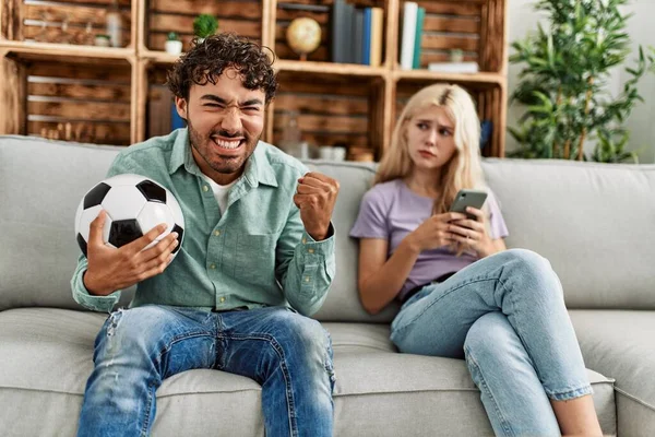Adam mutlu bir şekilde futbol maçı izliyor ve kız arkadaşı evde akıllı telefon kullanıyor..