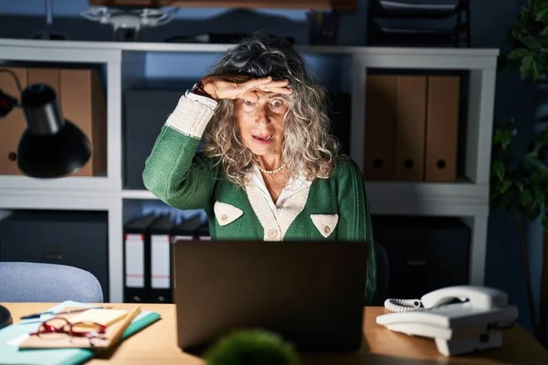 Middle age woman working at night using computer laptop very happy and smiling looking far away with hand over head. searching concept.