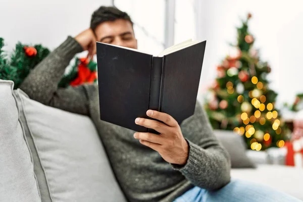 Young Hispanic Man Reading Book Sitting Sofa Christmas Tree Home — Stok fotoğraf
