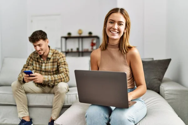 他的年轻夫妇笑着高兴地坐在沙发上使用笔记本电脑和智能手机 — 图库照片