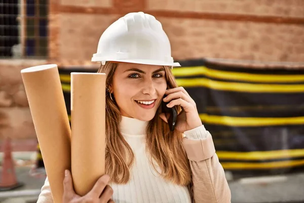 Young blonde woman holding blueprints talking on the smartphone at street