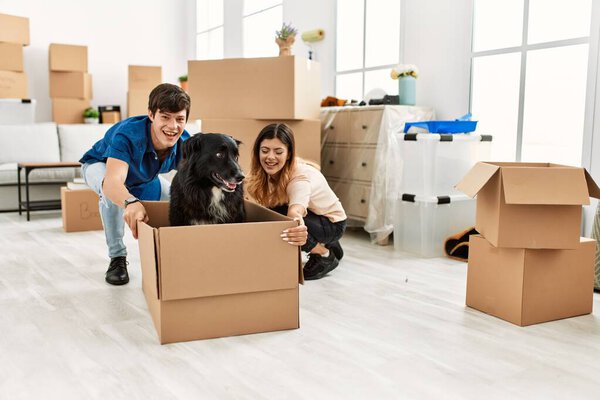 Молодая кавказская пара улыбается счастливой транспортировке собаки в картонную коробку в качестве автомобиля в новом доме.