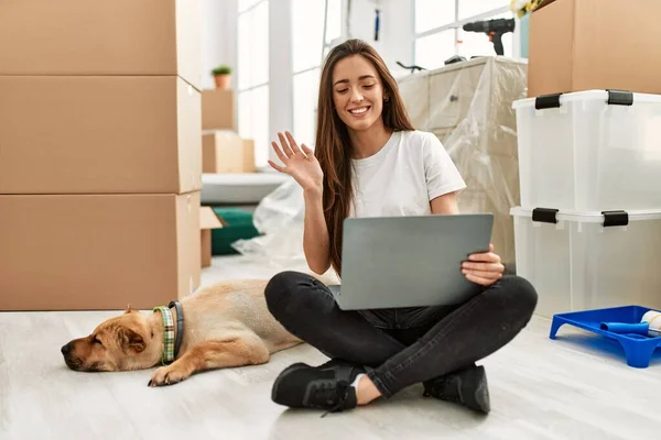 Young hispanic woman having video call sitting on floor with dog at new home