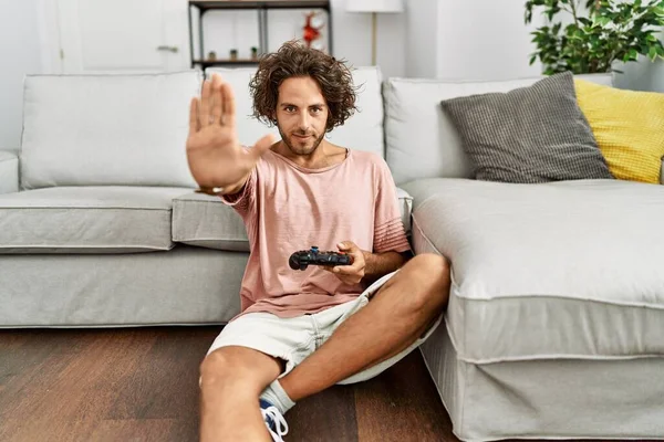 Young hispanic man playing video game holding controller at home with open hand doing stop sign with serious and confident expression, defense gesture