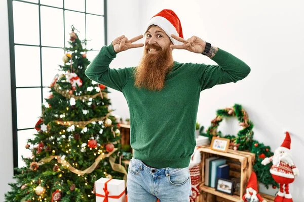 크리스마스 모자를 크리스마스 트리를 턱수염을 두르고 손가락을 얼굴에 평화의 상징을 — 스톡 사진