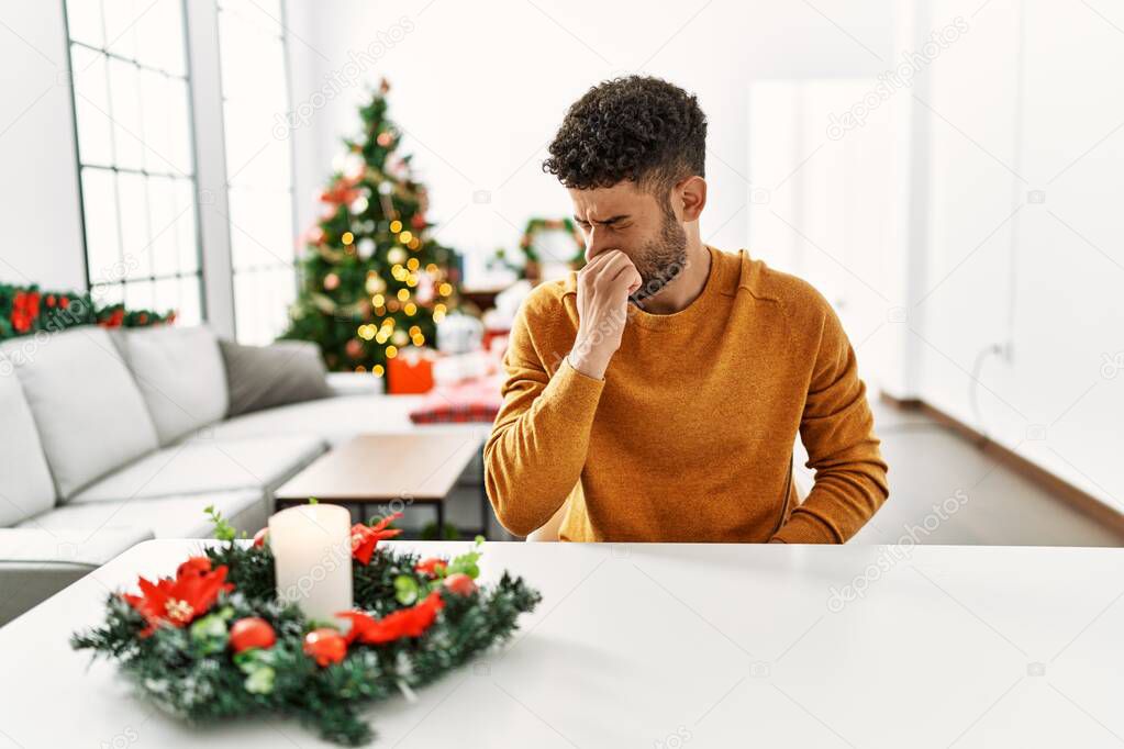 Arab young man sitting on the table by christmas tree smelling something stinky and disgusting, intolerable smell, holding breath with fingers on nose. bad smell 
