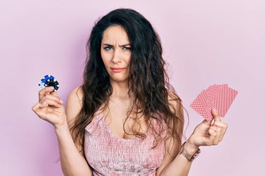Young hispanic woman playing poker holding casino chips and cards clueless and confused expression. doubt concept. 