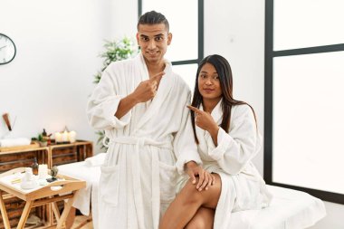 Havlu giymiş genç Latin bir çift, güzellik merkezinde el parmakla işaret ederek ciddi ve sakin bir yüz ifadesi sergiliyor. 
