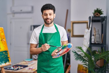 Genç Arap sanatçı gülümsüyor. Resim stüdyosunda fırça ve palet tutuyor.