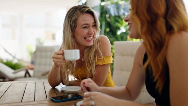 İki kadın evdeki terasta oturmuş kahve içiyor ve konuşuyor.