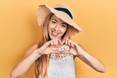 Yazlık şapka takan, aşık gülümseyen, elleri ile kalp sembolü yapan genç beyaz bir kadın. romantik kavram. 