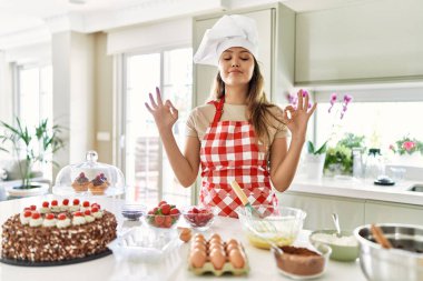 Güzel, genç, esmer pasta şefi kadın mutfakta hamur işleri pişiriyor gözleri kapalı gülümsüyor parmaklarıyla meditasyon hareketi yapıyor. yoga konsepti. 