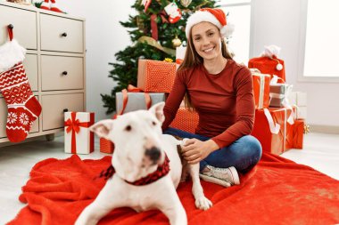 Beyaz bir kadın gülerek evde köpekle oturuyor. Noel ağacı yanında.