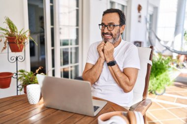Orta yaşlı bir adam evde bilgisayar kullanıyor. Gergin ve heyecanlı bir şekilde gülüyor. Elleri çenesinde yan tarafa bakıyor. 