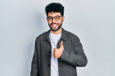 Sakallı, gözlüklü genç bir Arap, elleriyle mutlu bir jest yapıyor. Kameraya bakıp başarıyı gösteren ifade. 