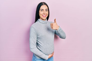 Mavi gözlü güzel bir kadın bebek bekliyor, hamile karnına dokunuyor mutlu bir el hareketi yapıyor. Kameraya bakıp başarıyı gösteren ifade. 