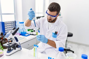 Laboratuvarda pipet ve test tüpü kullanarak bilim adamı üniforması giyen İspanyol genç bir adam.