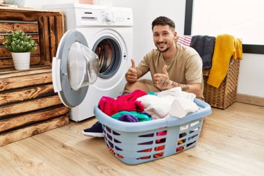 Young handsome man putting dirty laundry into washing machine success sign doing positive gesture with hand, thumbs up smiling and happy. cheerful expression and winner gesture. 
