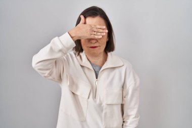 Middle age hispanic woman standing over isolated background covering eyes with hand, looking serious and sad. sightless, hiding and rejection concept 