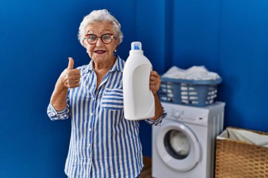 Beyaz saçlı, elinde deterjan şişesi olan yaşlı bir kadın mutlu ve pozitif gülümsüyor, başparmağıyla mükemmel ve onay işareti yapıyor. 
