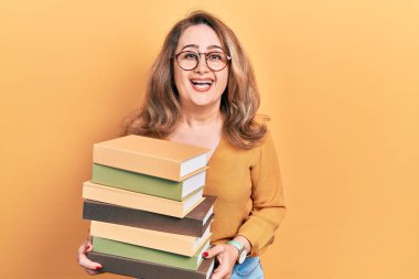 Middle age caucasian woman holding a pile of books smiling and laughing hard out loud because funny crazy joke. 