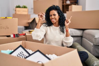 Middle age hispanic woman holding keys of new home smiling happy pointing with hand and finger to the side 