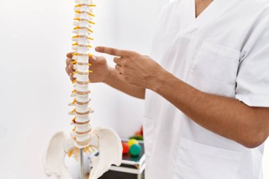 Fizyoterapist üniforması giyen genç Arap adam klinikte parmak omuza sütun gösteriyor.