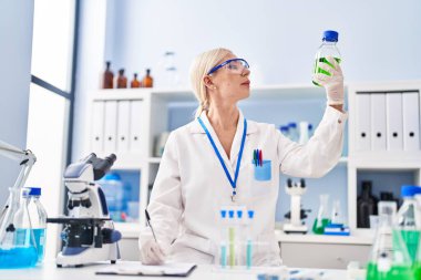 Laboratuvarda elinde şişe tutan bilim adamı üniforması giyen genç sarışın kadın.