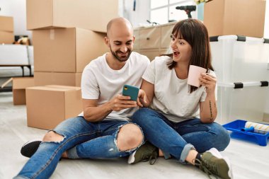 Genç İspanyol çift yeni evinde akıllı telefon kullanarak mutlu bir şekilde gülümsüyor..
