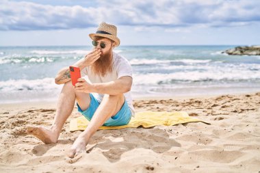 Genç kızıl saçlı adam sahilde havlunun üzerinde otururken akıllı telefonun yanında selfie çekiyor..