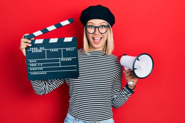 Beautiful blonde woman holding video film clapboard and megaphone celebrating crazy and amazed for success with open eyes screaming excited. 