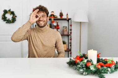 Noel süslemesi masasında oturan sakallı yakışıklı genç adam el hareketleriyle gülümsüyor ve mutlu yüzüyle parmaklarının arasından bakıyor.. 