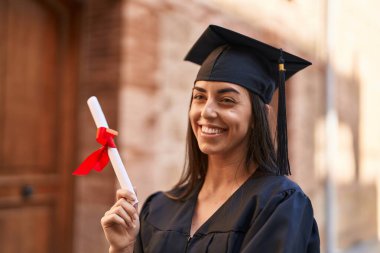 Young hispanic woman wearing graduated uniform holding diploma at university