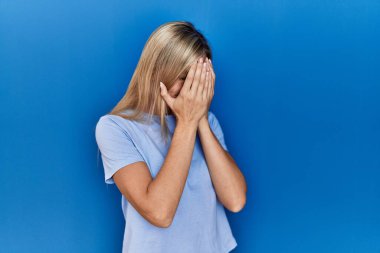 Güzel sarışın kadın mavi arka planda sıradan bir tişört giyiyor. Ağlarken suratını ellerle kapatıyor. depresyon kavramı. 
