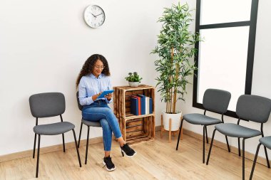 Bekleme odasında sandalyede oturan dokunmatik ped kullanan genç Latin kadın.