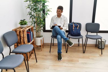 Young hispanic man waiting filling documents on clipboard at waiting room