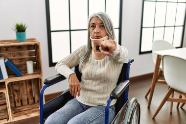 Orta yaşlı, gri saçlı, tekerlekli sandalyede oturan, tasmalı, kamerayı işaret eden, kendine güvenen, ciddi görünen bir kadın. 