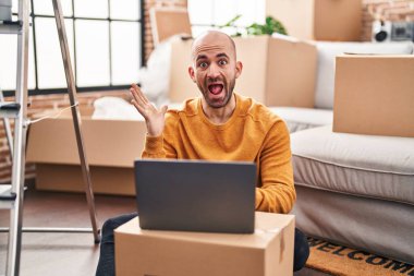Young bald man with beard moving to a new house using laptop celebrating victory with happy smile and winner expression with raised hands 