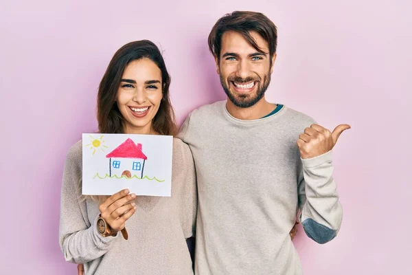 Young Hispanic Couple Holding House Draw Pointing Thumb Side Smiling — Stockfoto