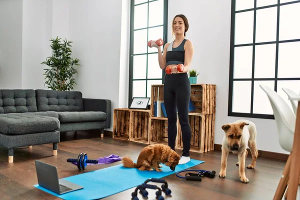 Young hispanic woman smiling confident having online traininig with dogs using dumbbells at home
