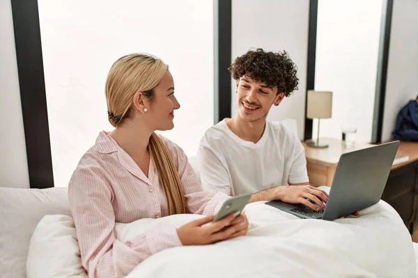 年轻漂亮的夫妇坐在家里的床上 用笔记本电脑和智能手机 — 图库照片